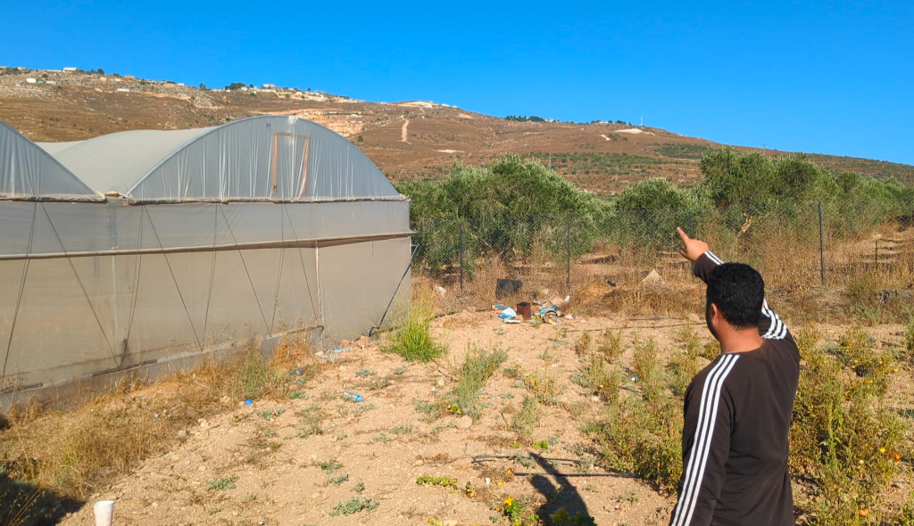 A man points to the houses at the top of a hill behind greenhouses